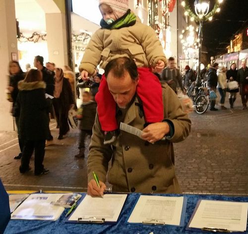 '图7：一位德国男士顶着孩子认真的签名支持法轮功'